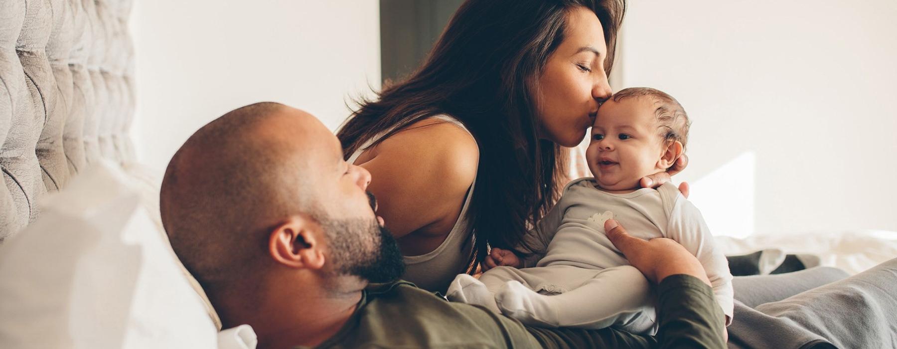 mother kisses her infant while father lies in bed and holds him